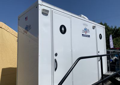 Exterior view of a clean, white luxury restroom trailer with gender-specific signage and LED porch lighting for evening corporate events.