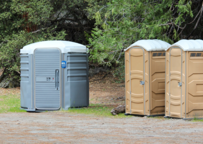 A large, circular grey handicap-accessible portable toilet placed next to two tan standard units in a natural, wooded park setting.